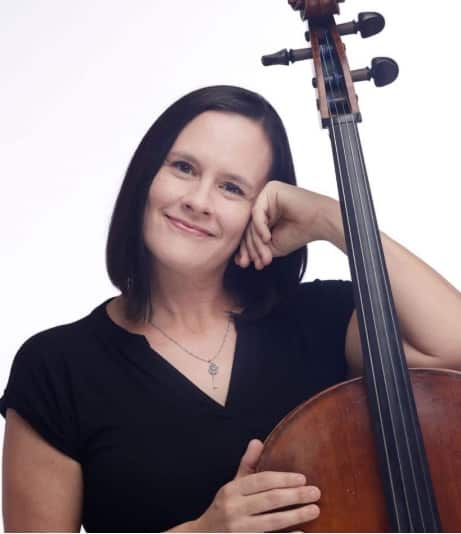 A young woman with dark hair wearing a black shirt holds a cello and smiles at the camera against a white background.