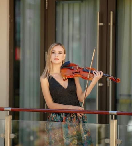 A young woman playing violin on a balcony, wearing a black sleeveless top and patterned skirt, with a blurred urban background.