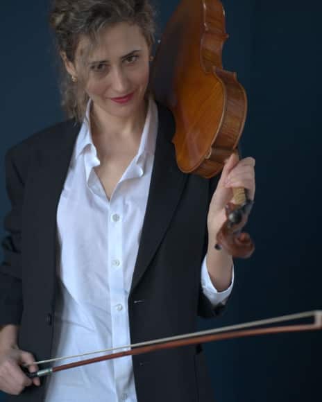 A woman in a black blazer and white shirt holds a violin and bow against a dark blue background.