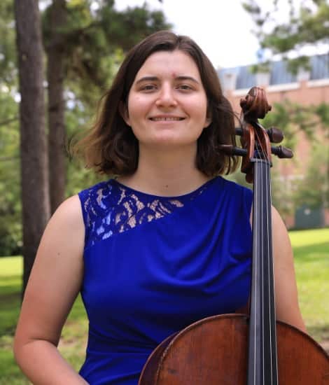 A young woman in a blue lace dress holding a cello, smiling at the camera outdoors with trees and green grass in the background.