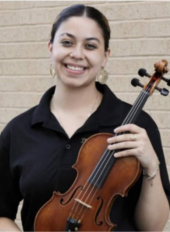 A smiling young woman with dark hair wearing a black polo shirt holds a violin while posing in front of a brick wall.