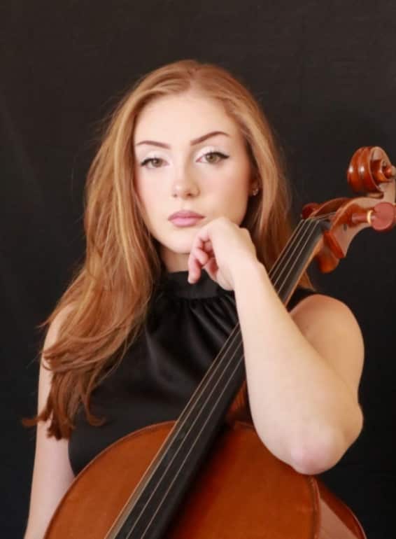 A young woman with long reddish-brown hair wearing a black sleeveless top, posing thoughtfully with her hand near her face while holding a cello against her shoulder against a dark background.
