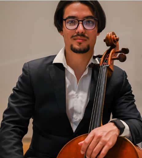 A man wearing glasses and a dark blazer with white shirt holds a cello, positioned against a neutral beige background.