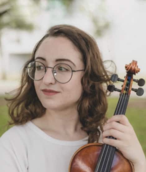 A young woman with shoulder-length brown hair and round glasses holds a violin while looking toward the camera outdoors.