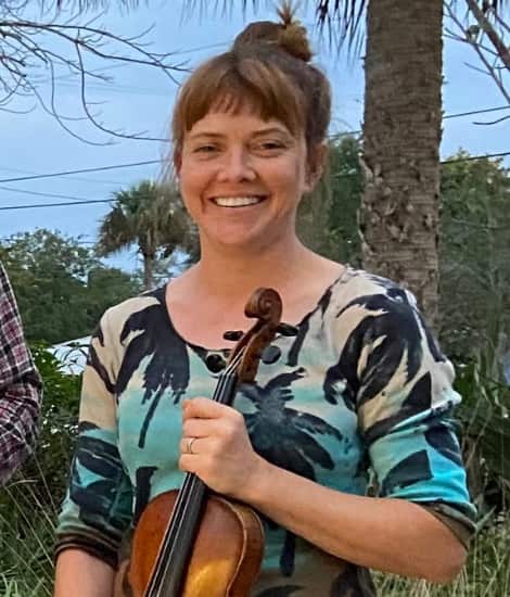 A smiling woman with brown hair in a bun holds a violin while standing outdoors with palm trees in the background.