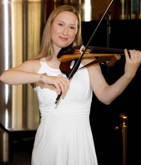 A woman in a white halter dress playing a violin, smiling at the camera in an elegant interior setting with a piano visible in the background.