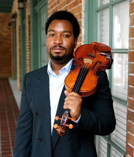 A man in a dark double-breasted blazer and white shirt holds a violin over his shoulder while posing in front of a brick building with green-trimmed windows.