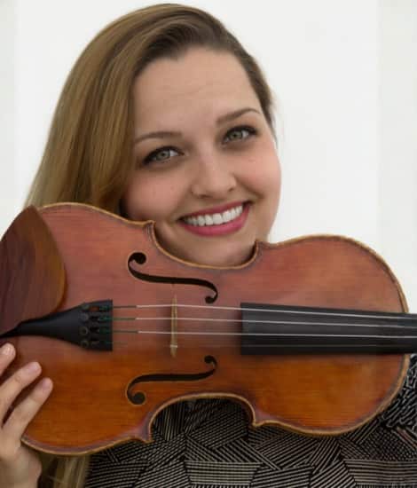 A smiling woman with long brown hair holding a violin against her shoulder, wearing a dark patterned top against a white background.