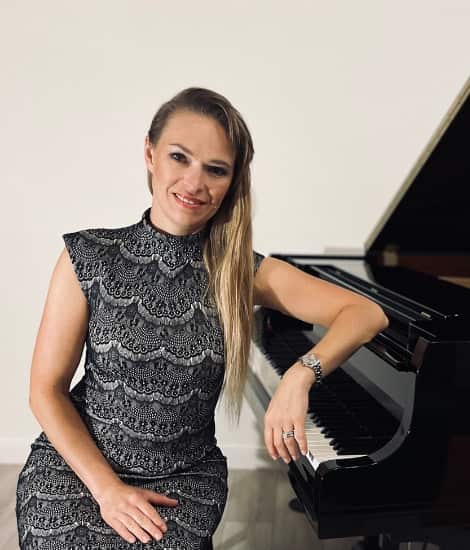 A woman with long blonde hair smiling at the camera while sitting at a grand piano, wearing a black lace sleeveless dress.