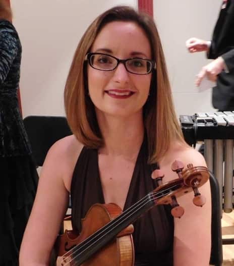 A woman with reddish-brown hair and glasses smiles at the camera while holding a violin. She is wearing a dark halter-style top and appears to be at an indoor venue or concert setting.