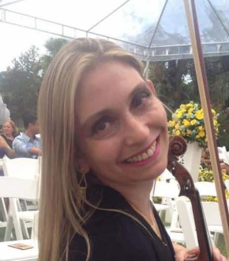 A smiling woman with long blonde hair wearing a black top and hoop earrings at an outdoor event under a clear tent, with yellow flowers and white chairs visible in the background.