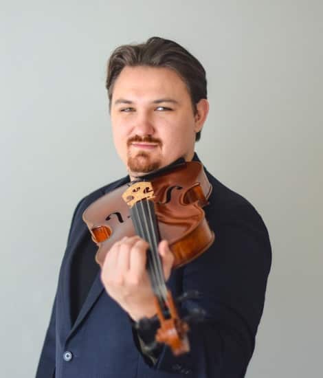 A man with dark hair and a goatee wearing a dark blue shirt holds a violin at an angle across his body against a neutral gray background.