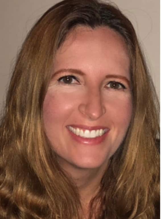 Professional headshot of a woman with shoulder-length brown hair, smiling at the camera against a neutral beige background.