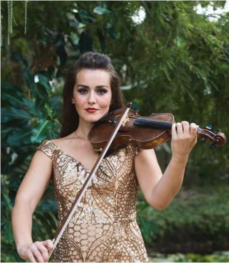 Woman in a gold patterned dress playing violin outdoors with green foliage in the background.