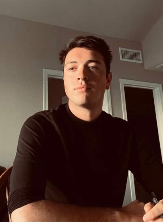 A man with dark brown hair wearing a black long-sleeved shirt, photographed indoors against a white wall with doors and a ceiling vent visible in the background.