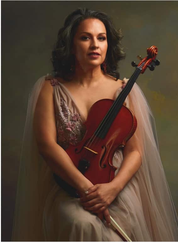 A woman with dark hair wearing an elegant beige and burgundy draped gown holds a deep red violin while gazing thoughtfully to the side against a muted green background.
