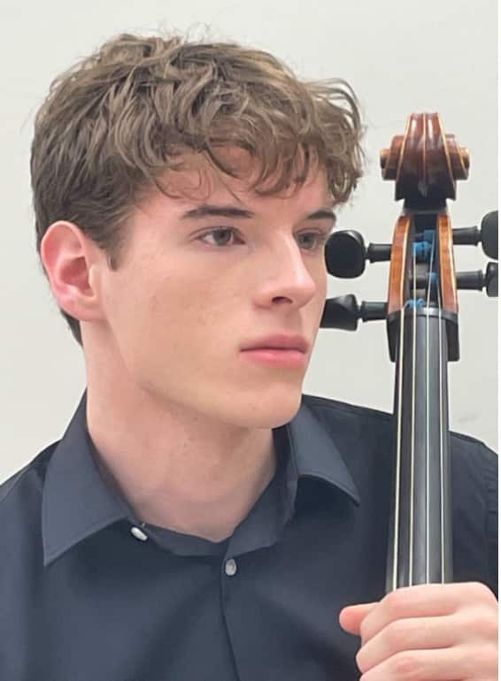 A young musician holding a cello, posing for a portrait against a white background while wearing a dark blue shirt.