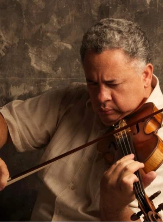 A middle-aged man with gray hair playing a violin, wearing a beige shirt, photographed against a textured brown background.