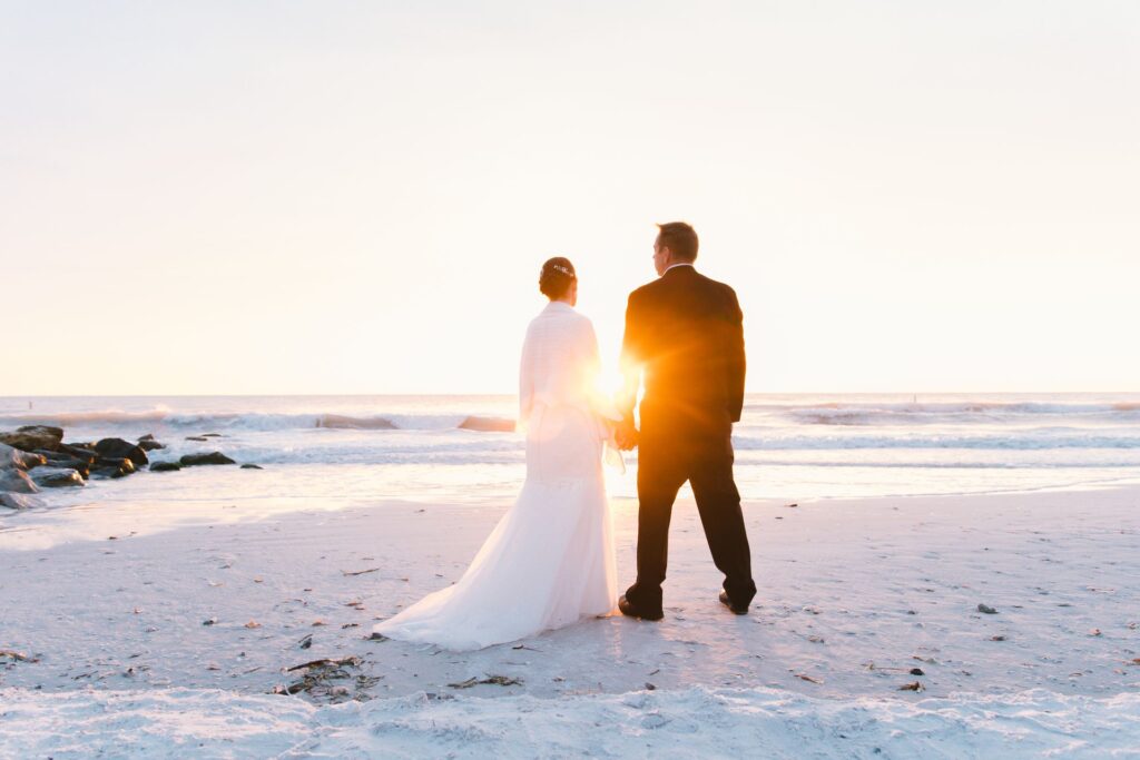 A bride and groom stand together on a beach at sunset, holding hands and gazing at the ocean horizon. The groom wears a dark suit while the bride wears a white wedding dress with a flowing train. Golden sunlight illuminates them from behind as waves crash on the shore.