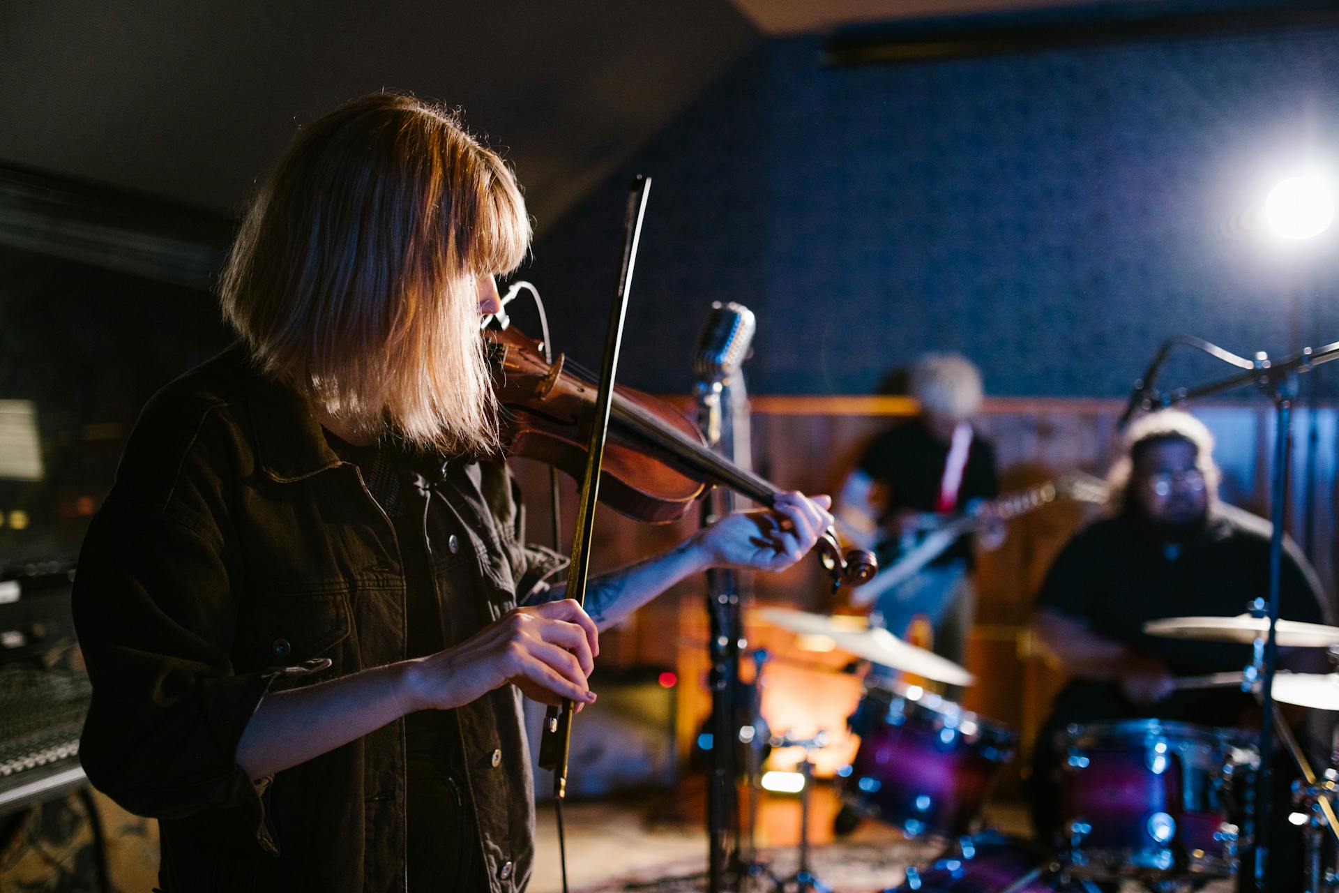 A violinist with long blonde hair performs on stage under stage lighting, with band members and drums visible in the blurred background during a live musical performance.