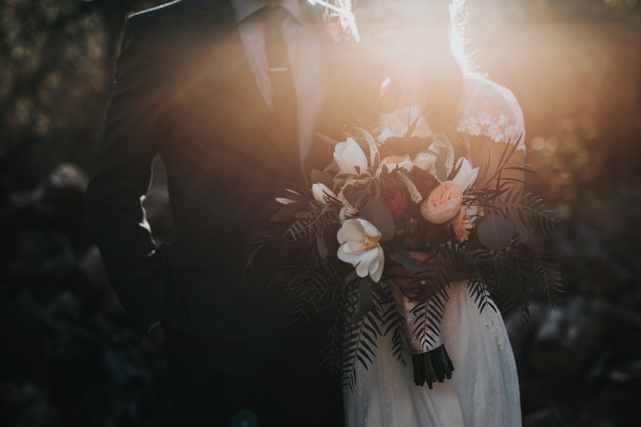 A bride and groom stand together in golden sunlight, with the bride holding a bouquet of white and peach flowers with fern greenery.
